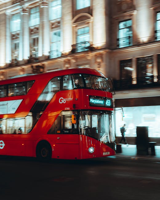 At night, a line of red double-decker buses is stopped on a city street in Marylebone, London. The closest bus displays a digital sign reading 'Marylebone' and route number 453. Behind the buses, there is a modern, curved building illuminated with blue lighting, and additional vehicles are visible with headlights on, creating a bright contrast against the dark sky. The scene captures the urban environment typical of a busy London area, with the buses awaiting passenger boarding or preparing for departure, and some individuals can be seen inside the buses through the windows. The overall setting reflects a typical evening at a transport hub supporting house removals, furniture transport, and moving logistics, with the buses possibly involved in home relocation services or transporting clients to or from a move. Man with Van Marylebone's service is implied by the context, although not directly depicted. The street is lit by streetlights, and the pavement runs along the left edge of the image.