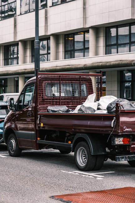 A dark red compact pickup truck parked on a city street in front of a modern office building with large glass windows. The truck's open cargo bed is filled with various items, including cardboard boxes, large plastic-wrapped packages, and soft furnishings, indicating a home relocation or furniture transport process. The cargo includes wrapped furniture pieces, some covered with protective fabric or plastic to prevent damage during loading or transport, with items positioned securely inside the bed. A metal grid barrier is installed behind the cab, and the vehicle is situated close to a nearby curb with a red tactile paving strip visible in the foreground. Surrounding the truck, other vehicles are parked along the street, and a street lamp pole is partially visible behind the truck, suggesting an urban environment. This scene reflects the logistics involved in house removals or clearance services, supported by a professional moving company like Man with Van Marylebone.