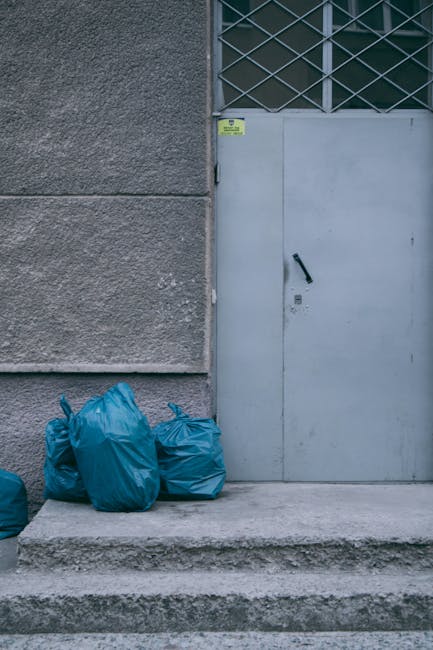 Several blue plastic bags filled with bulky waste items are placed on the stone steps outside a building entrance, adjacent to a grey metal door with a black handle and lock. The building's exterior wall is constructed of textured concrete, and there is a small yellow notice above the door. In the background, a metal grille is visible above the door. This scene depicts a common setup for home relocation or furniture transport, with waste awaiting removal or disposal as part of the packing and moving process. The environment is well-lit with natural light, and the bags are positioned on the pavement, ready for collection or loading into a vehicle. The image relates to professional removals services offered by Man with Van Marylebone, specialising in house and furniture removals, including bulky waste removal at Marylebone flats.