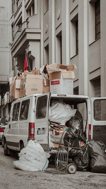 A dark red compact pickup truck parked on a city street in front of a modern office building with large glass windows. The truck's open cargo bed is filled with various items, including cardboard boxes, large plastic-wrapped packages, and soft furnishings, indicating a home relocation or furniture transport process. The cargo includes wrapped furniture pieces, some covered with protective fabric or plastic to prevent damage during loading or transport, with items positioned securely inside the bed. A metal grid barrier is installed behind the cab, and the vehicle is situated close to a nearby curb with a red tactile paving strip visible in the foreground. Surrounding the truck, other vehicles are parked along the street, and a street lamp pole is partially visible behind the truck, suggesting an urban environment. This scene reflects the logistics involved in house removals or clearance services, supported by a professional moving company like Man with Van Marylebone.