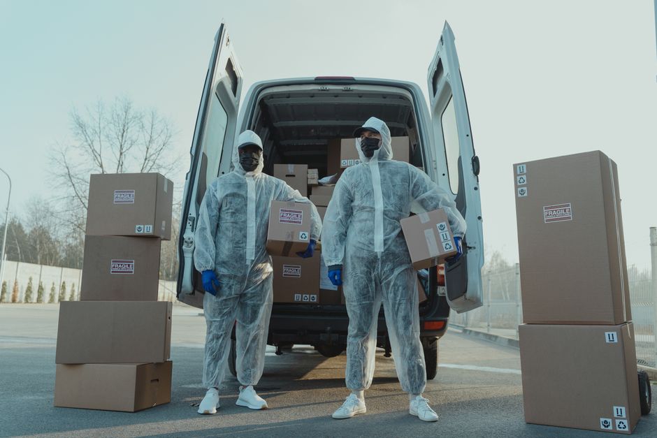 Two moving staff members dressed in full protective suits, face masks, and gloves are standing outside a white van with its rear doors open, loading and unloading cardboard boxes sealed with packing t