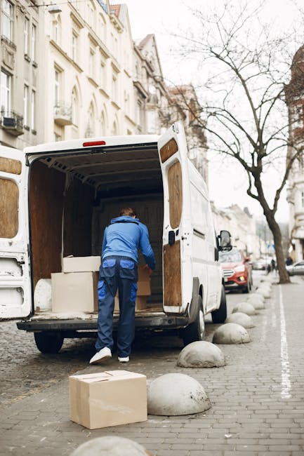 Two moving staff members dressed in full protective suits, face masks, and gloves are standing outside a white van with its rear doors open, loading and unloading cardboard boxes sealed with packing t
