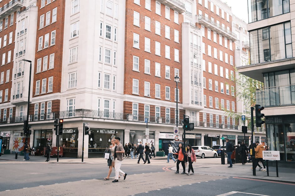 A busy urban street scene in Marylebone, showing a multi-storey building with a combination of red brick and white facade, housing various retail outlets on the ground floor, including a bakery and cafes. Pedestrians are crossing the road at the crosswalk, some carrying shopping bags, while others are walking along the pavement. Several street traffic lights and lampposts are visible, with a few cars waiting at the intersection. To the right, a modern building with glass balconies is partially visible. In the foreground, a group of people is engaged in a home relocation or moving process, with cardboard boxes, packing materials, and a trolley positioned nearby, indicating ongoing packing and furniture transport activities consistent with house removals by Man with Van Marylebone.
