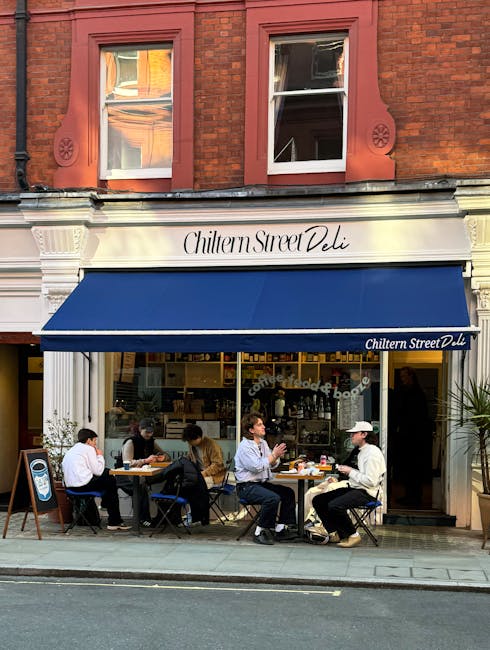 Photograph of the exterior of Chiltern Street Deli, a restaurant situated on the ground floor of a red brick building with two upper windows. The storefront features a large beige sign with the shop's name in elegant black script, and a dark blue canopy extending over the outdoor seating area. In front of the deli, five people are seated at black metal tables with matching chairs, engaged in conversation and enjoying meals or drinks. The individuals are dressed casually, with some wearing jackets and masks, indicating recent or ongoing health precautions. The seating area is on the sidewalk, directly adjacent to the street, which has a single yellow line. The scene captures a typical urban setting focused on food service, with natural daylight illuminating the scene and a glimpse of the interior visible through the glass windows behind the patrons. The image emphasizes the inviting street-level atmosphere of a busy, popular food and beverage establishment, relevant to relocation, packing, and transport contexts as part of a house removal or moving service in Marylebone.