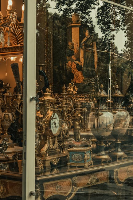 A view through a large glass window displaying an interior filled with antique decorative items including ornate brass candelabras, vintage clocks, and porcelain vases, indicating an antique shop in Marylebone. Outside, two movers dressed in work clothing are seen carefully handling large, fragile antique objects, preparing for transportation as part of a home relocation or furniture transport process. The scene captures the unloading or loading process, with the antiques positioned on a trolley or moving equipment, and the natural outdoor lighting highlighting the reflective glass surface. This image illustrates the careful handling and packing of valuable antiques during a professional removals service, such as those offered by Man with Van Marylebone, emphasizing expertise in delicate furniture transport and packing in moving logistics.