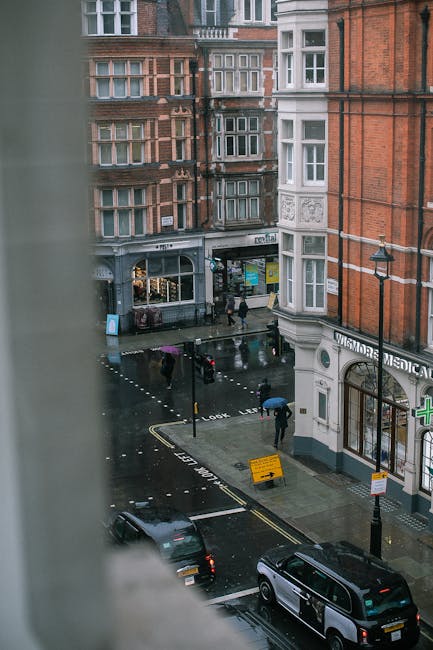 View of a rainy street scene in Marylebone from a higher window, showing a black van parked on the wet pavement and several pedestrians holding umbrellas walking past a building with a ground-floor pharmacy and a clothing store. The street features road markings, including a yellow barrier and a pedestrian crossing with white dotted lines, while the surrounding buildings display red brick and white facade details, typical of the area. Man with Van Marylebone's removals service would coordinate vehicle access and loading from this area during house relocation or furniture transport, with the wet conditions and urban environment requiring careful planning for a smooth packing and moving process.