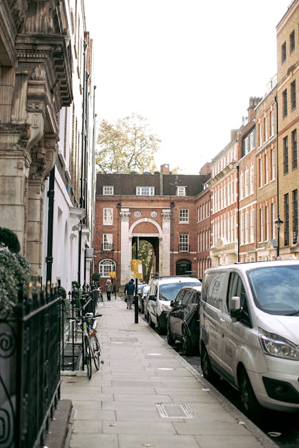 A narrow urban street scene shows a row of parked vehicles, including a white van, along the pavement outside residential buildings featuring brick facades and traditional architectural details. On the left, a bicycle is secured to a black wrought iron fence, and a few pedestrians are visible in the distance near a large arched gateway set within a red-brick building. The pavement is made of grey stone slabs, and the street is lined with trees and lampposts. Overhead, the sky is clear with some foliage from a tree visible at the top of the image. This scene depicts the typical environment for home relocation or furniture transport, illustrating the loading process as part of a professional removals service, such as those offered by Man with Van Marylebone.