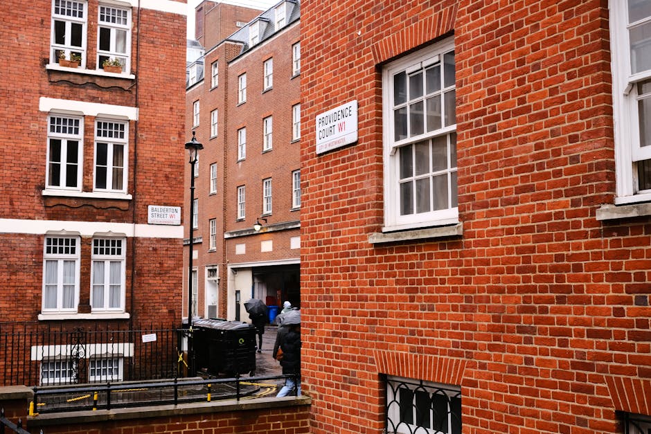 Photograph showing the narrow exterior alleyway between red-brick residential buildings in Marylebone, London, with visible white-framed windows. A worker from Man with Van Marylebone, a company specializing in removals, is seen loading or unloading a large black waste disposal bin situated on the pavement near the building entrance. The alley contains a black metal fence and a yellow and black barrier, with a streetlight positioned along the corner of the buildings. Two men are engaged in moving activities, one wearing a grey jacket and the other partially visible, both involved in the home relocation or furniture transport process. Signs on the buildings indicate the streets as Providence Court and Balderton Street, providing context for the location of the last-minute booking services offered by Man with Van Marylebone. The lighting suggests daytime, with natural light illuminating the scene, capturing the logistics involved in packing and moving within an urban setting.