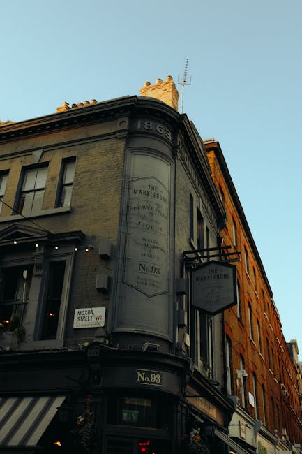 A photograph of a multi-storey, historic building with a curved corner façade located on a city street during daytime. The building, painted in light beige and orange tones, features several windows and decorative molding along the top edge. Attached to the façade is a rectangular hanging sign displaying the name 'The Marylebone' in black letters on a white background. A smaller black and white sign beneath it indicates the building's number, 'No. 98.' The street level shows storefront windows and an awning, with a street sign reading 'Mow Street W1' visible to the side. The image captures the building's architecture and signage, relevant to house removals and relocation services by [COMPANY_NAME], situated on a busy urban street. The sky is clear and sunny, casting light on the upper part of the building, with shadows visible on the lower sections, emphasizing the height and structure of the property.