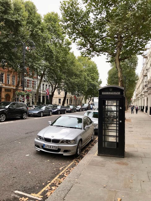 A silver BMW car parked partially on the curb and pavement alongside a black traditional British phone booth on a tree-lined street in Marylebone, with multiple parked vehicles further down the road. The scene is set in daytime with natural lighting, showing the street extending into the distance with pedestrians walking along the sidewalk. In the background, there are residential and commercial buildings with decorative facades, some with Union Jack flags hanging outside. This image depicts a typical urban setting relevant to house removals and furniture transport services, with visible parking and loading considerations that [COMPANY_NAME] might encounter during home relocation or moving logistics in the W1U and W1G postcode areas.