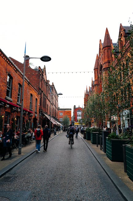 A street scene in Marylebone, London, showing pedestrians walking along the pavement and a cyclist riding down the cobblestone road during daytime. On the left side, there are outdoor seating areas with tables and chairs in front of cafes or restaurants, some of which are illuminated with string lights. The buildings are constructed with red brick, featuring large windows and decorative facades, and are lined with street lamps attached to poles. On the right side, there are tall, historic red brick buildings with pointed arches and gabled roofs, partially covered in green ivy or climbing plants. Several small trees and large planter boxes with greenery are positioned along the sidewalk. The scene captures a typical busy urban environment suitable for house removals or moving services, emphasizing the importance of careful furniture transport and loading logistics with the potential for street permits. The sky is overcast, providing natural lighting for the street scene, which is relevant to the context of residential and commercial relocations managed by companies like Man with Van Marylebone.