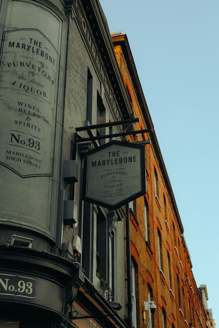 A street scene in Marylebone, London, showing pedestrians walking along the pavement and a cyclist riding down the cobblestone road during daytime. On the left side, there are outdoor seating areas with tables and chairs in front of cafes or restaurants, some of which are illuminated with string lights. The buildings are constructed with red brick, featuring large windows and decorative facades, and are lined with street lamps attached to poles. On the right side, there are tall, historic red brick buildings with pointed arches and gabled roofs, partially covered in green ivy or climbing plants. Several small trees and large planter boxes with greenery are positioned along the sidewalk. The scene captures a typical busy urban environment suitable for house removals or moving services, emphasizing the importance of careful furniture transport and loading logistics with the potential for street permits. The sky is overcast, providing natural lighting for the street scene, which is relevant to the context of residential and commercial relocations managed by companies like Man with Van Marylebone.