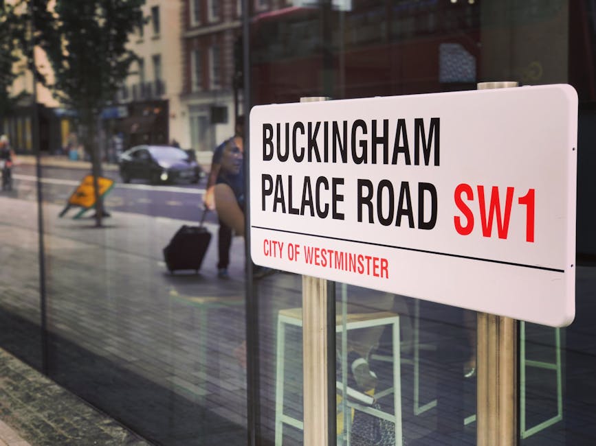 A white street sign displaying 'BUCKINGHAM PALACE ROAD SW1' with black capital letters and 'CITY OF WESTMINSTER' in red, mounted on metallic posts, positioned outside a modern building with reflective