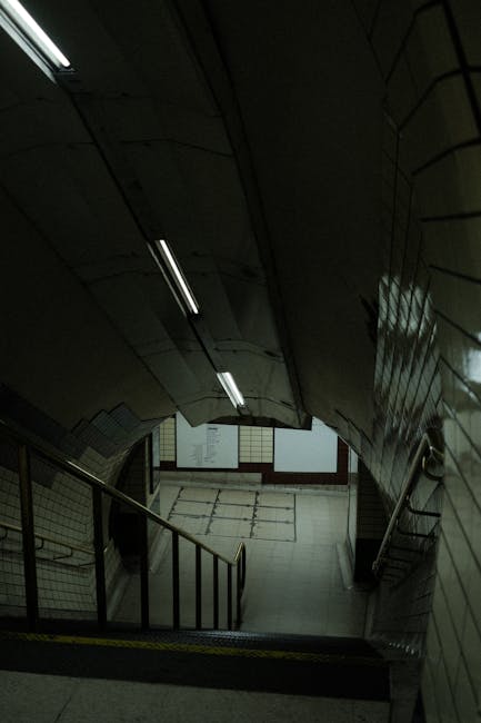 A dimly lit staircase inside a building, viewed from the top looking down, with metal handrails on both sides and tiled walls. The stairs are made of concrete with painted edges, and there is a small landing at the bottom. Fluorescent lighting runs along the ceiling, providing illumination for the passage. The environment appears enclosed and secure, typical of an internal stairwell used during home relocation or furniture transport processes. This setting is consistent with urban building interiors where household items are moved by professional removal services such as Man with Van Marylebone, especially when navigating narrow or stair-accessible properties for house removals and pack-and-move operations.