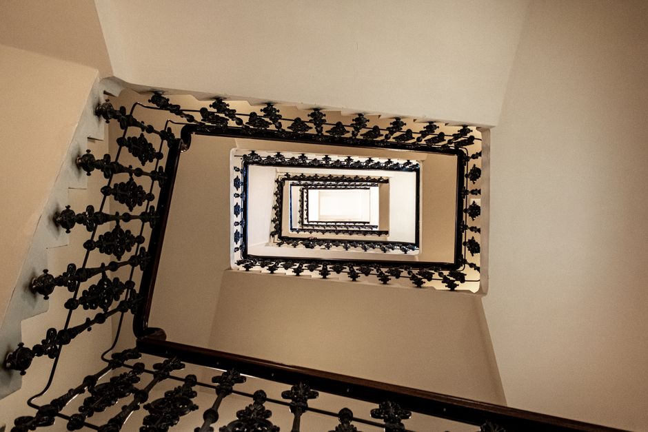 A dimly lit staircase inside a building, viewed from the top looking down, with metal handrails on both sides and tiled walls. The stairs are made of concrete with painted edges, and there is a small landing at the bottom. Fluorescent lighting runs along the ceiling, providing illumination for the passage. The environment appears enclosed and secure, typical of an internal stairwell used during home relocation or furniture transport processes. This setting is consistent with urban building interiors where household items are moved by professional removal services such as Man with Van Marylebone, especially when navigating narrow or stair-accessible properties for house removals and pack-and-move operations.